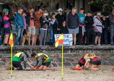 Two teams doing a puzzle on St Heliers beach while the crowd on the footpath watches and cheers them on